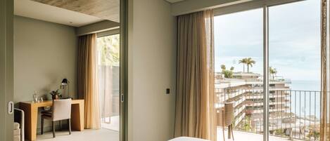 A hotel room with a large bed, a desk with a chair, and a view of the ocean and buildings.