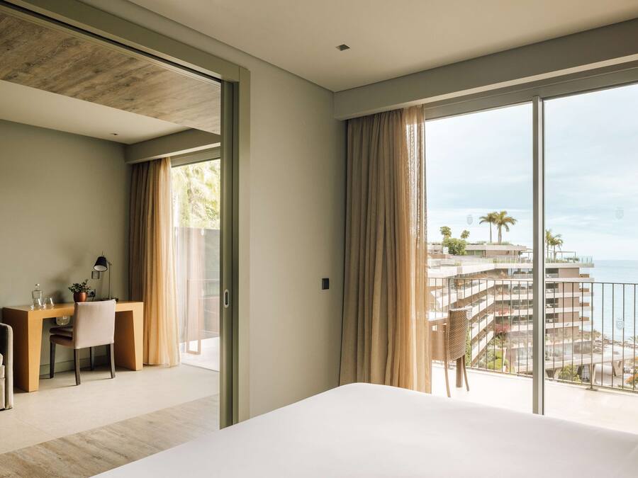 Een hotelkamer met een groot bed, een bureau met stoel, en uitzicht op zee en gebouwen.
