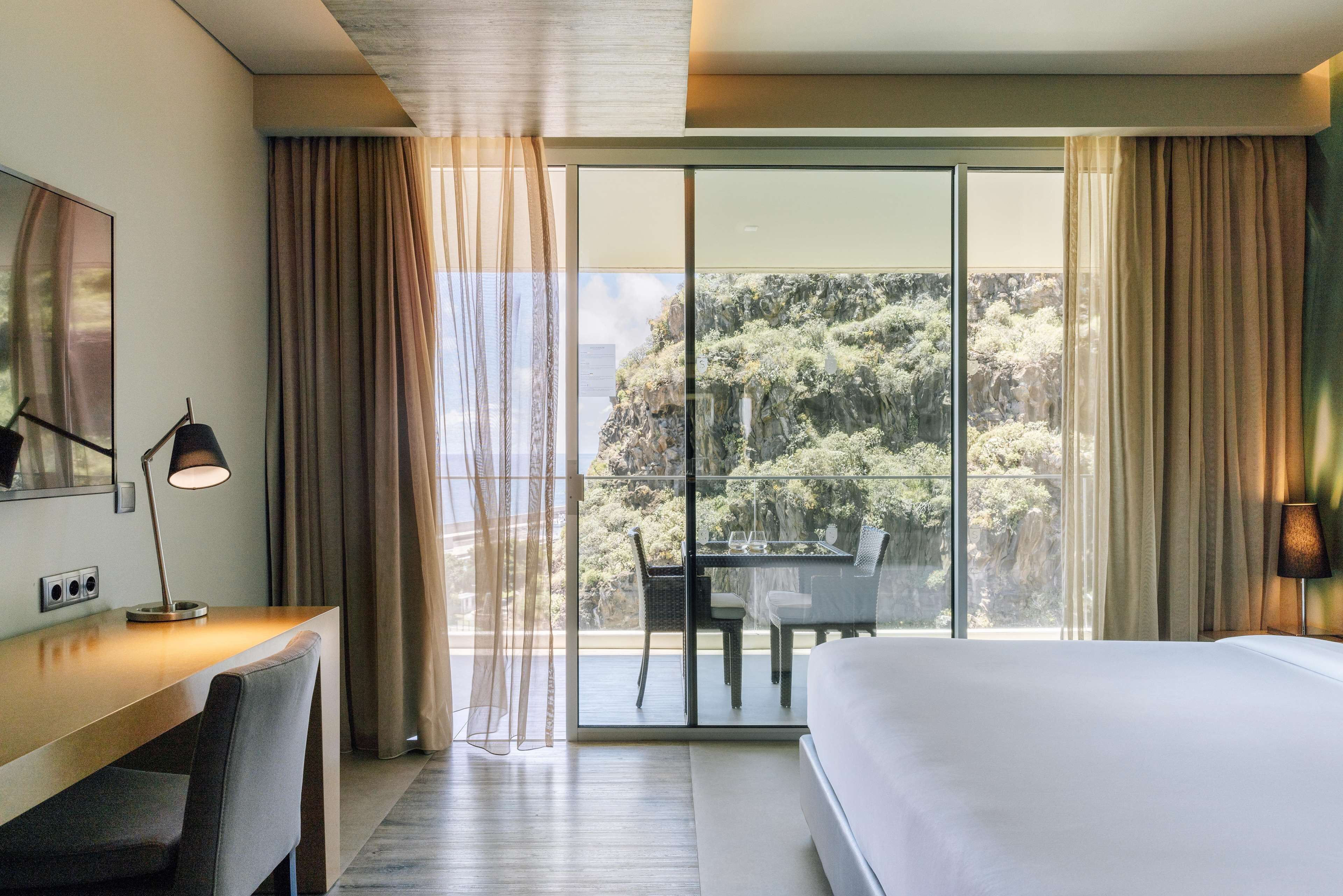 A hotel room with a large bed, a desk with a lamp, and a balcony with a table and chairs.