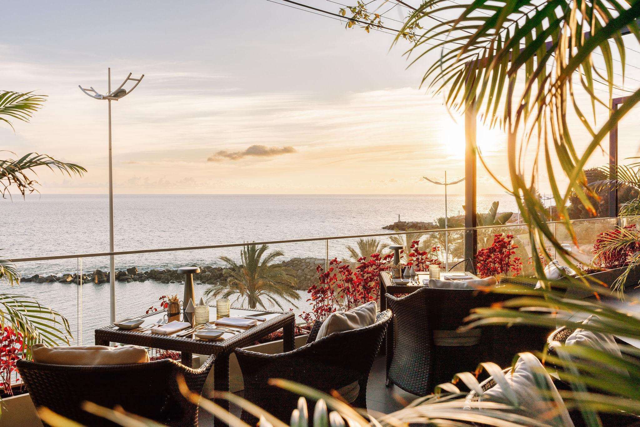 A balcony with wicker furniture, a table with a vase of red flowers, and a view of the sea.