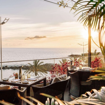 A balcony with wicker furniture, a table with a vase of red flowers, and a view of the sea.