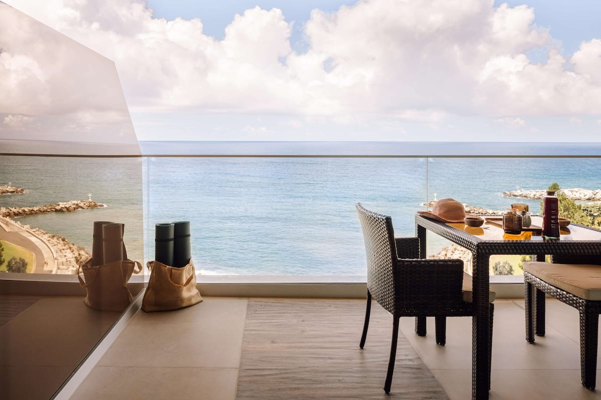 A balcony with a view of the sea, a wicker chair, a table with a hat and a drink, and a bag on the floor.