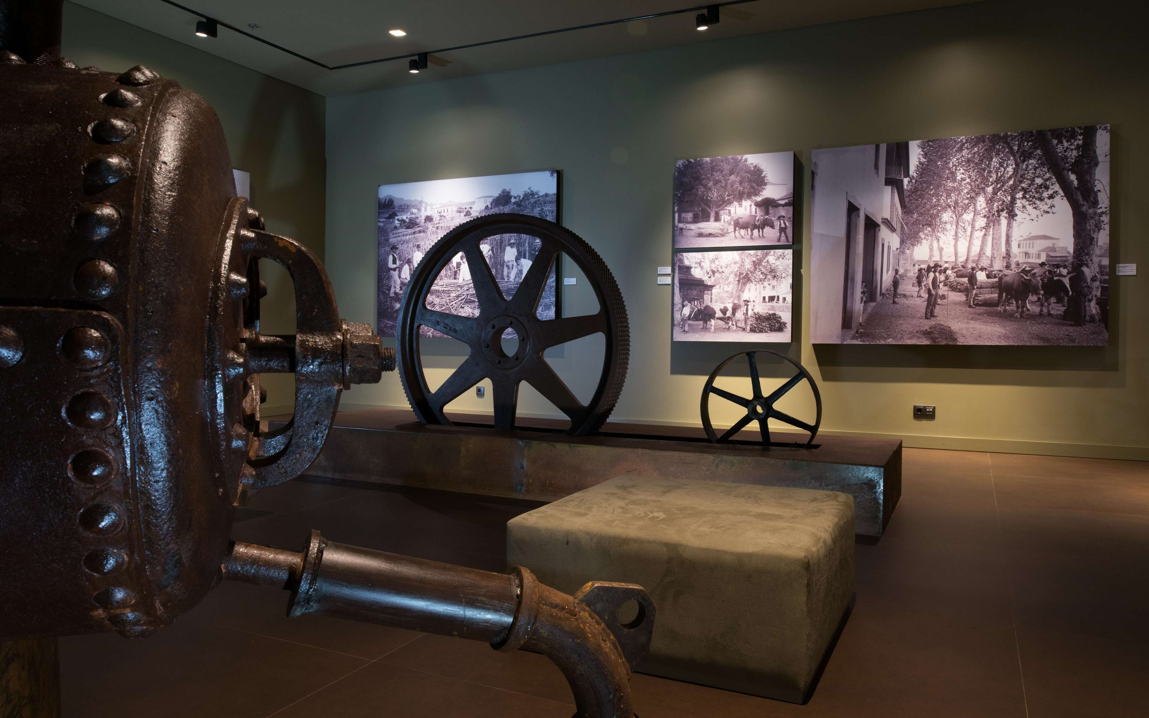 Una exposición de museo con maquinaria industrial, incluyendo ruedas grandes y tuberías, y fotografías históricas en la pared.