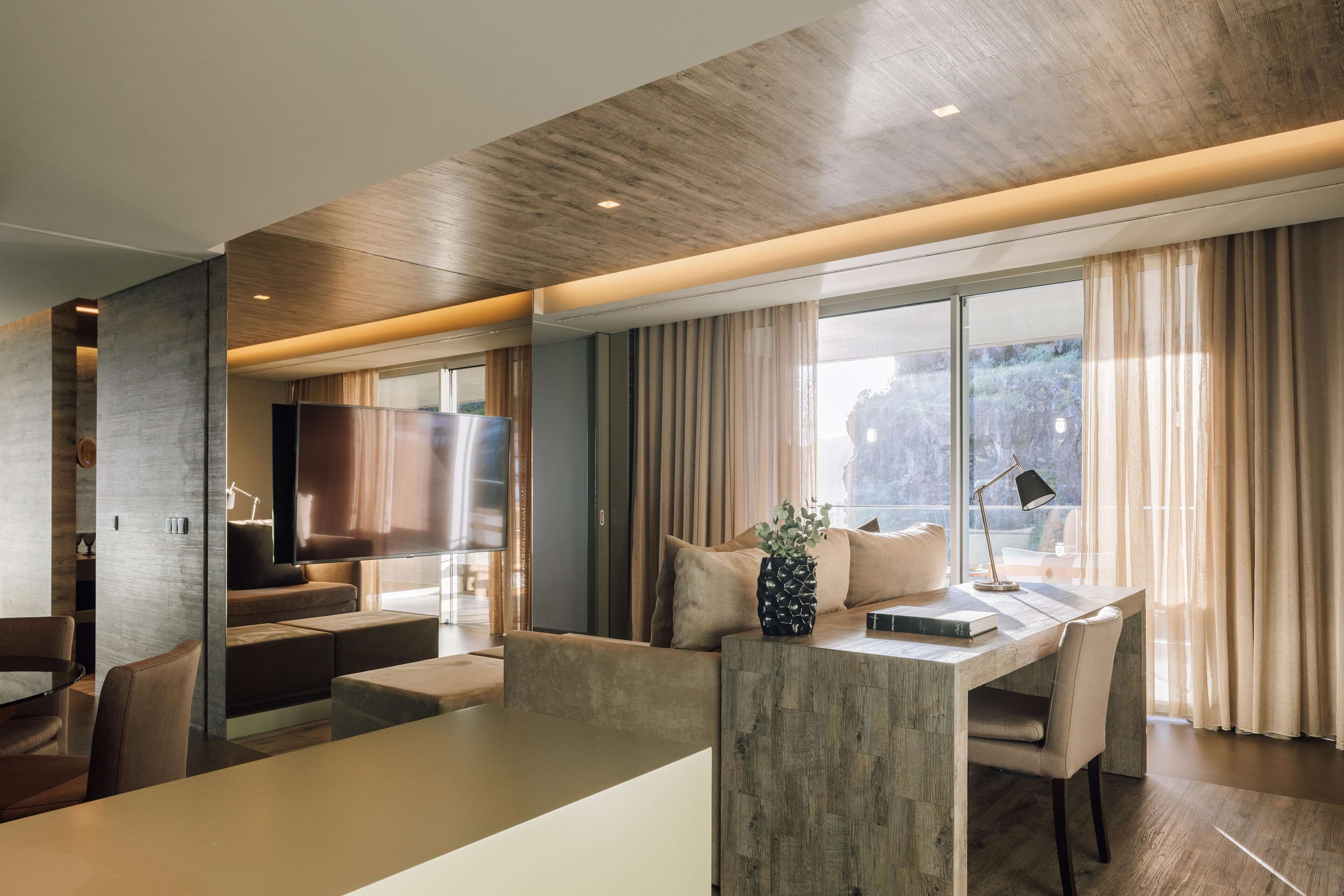 Uma sala de estar moderna com teto de madeira, uma janela grande com cortinas, um sofá, uma mesa de centro e uma escrivaninha com abajur.