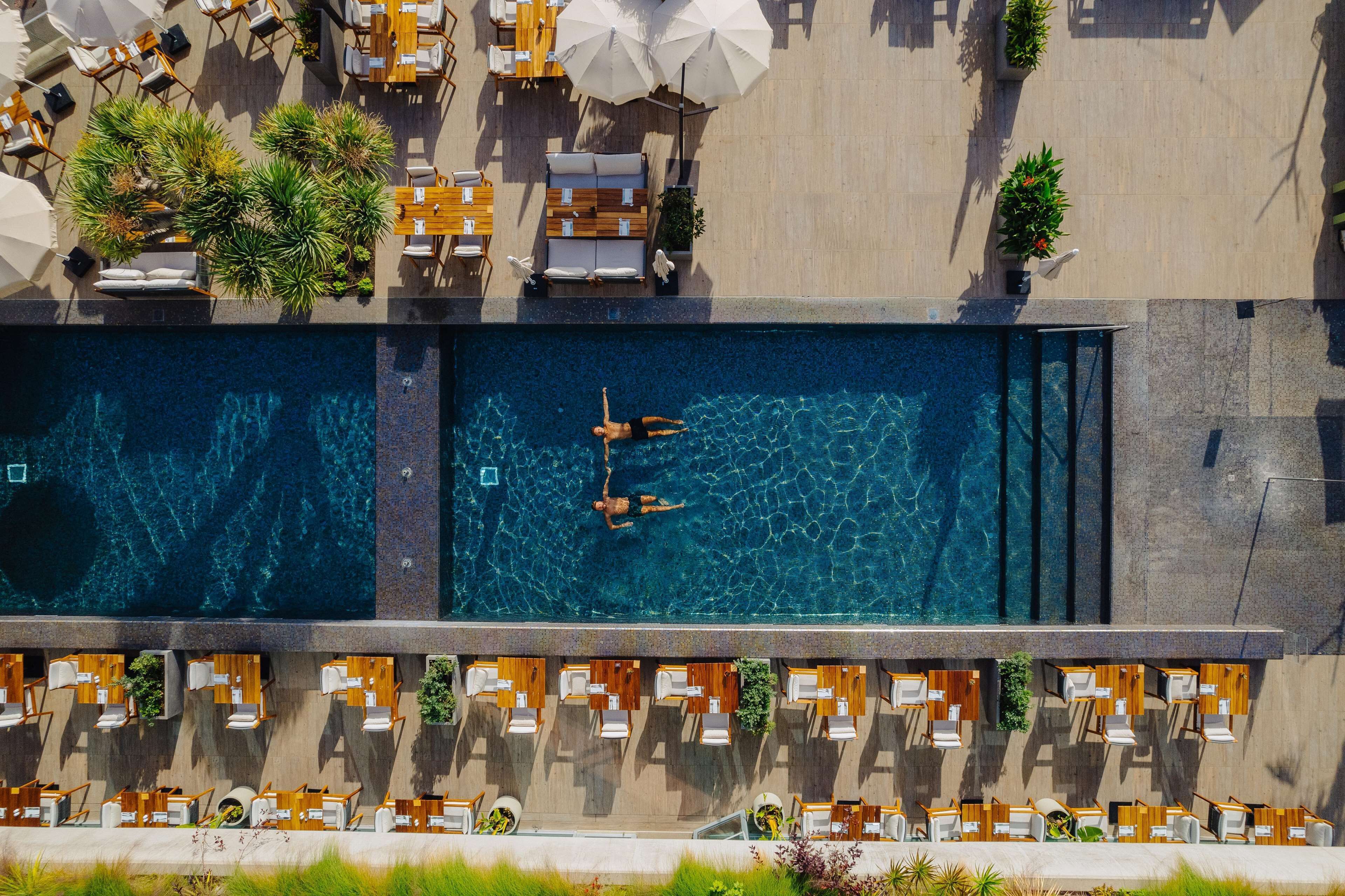 Vista aérea de una piscina en la azotea con dos personas nadando, rodeada de mobiliario de exterior y macetas con plantas.
