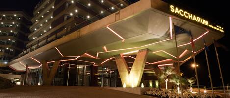 A modern hotel entrance with a prominent 'SACCHARUM' sign, a geometric roof design, and a landscaped area with plants.