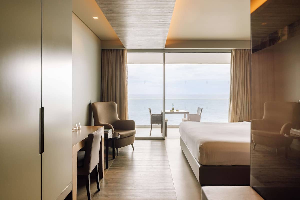 A modern hotel room with a large bed, two armchairs, a dining table, and a view of the sea.