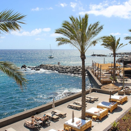 A beachfront resort with lounge chairs and umbrellas, a rocky shoreline, and a sailboat in the distance.