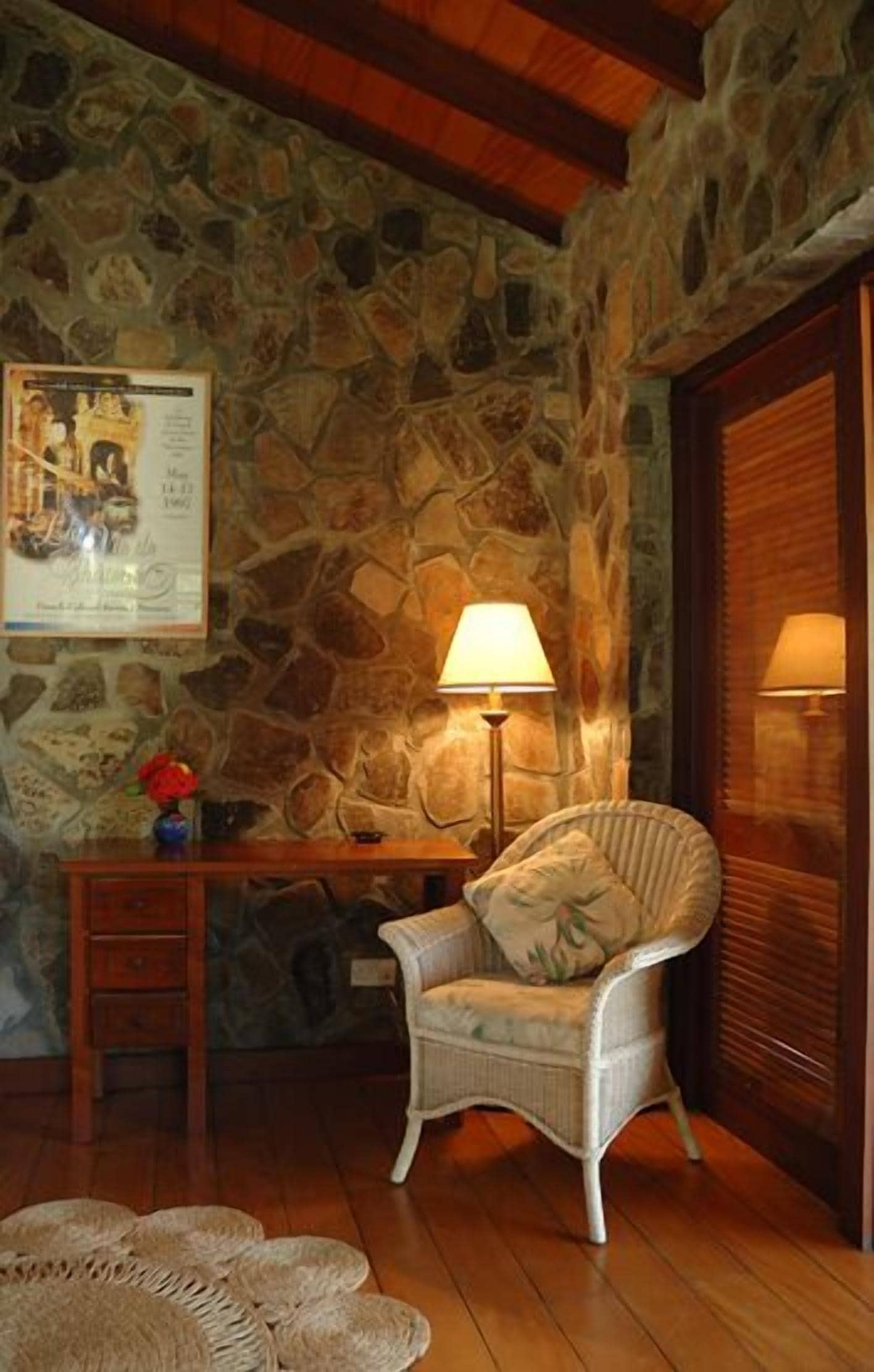 A cozy room with a stone wall, a wooden desk, a wicker chair, and two lamps.