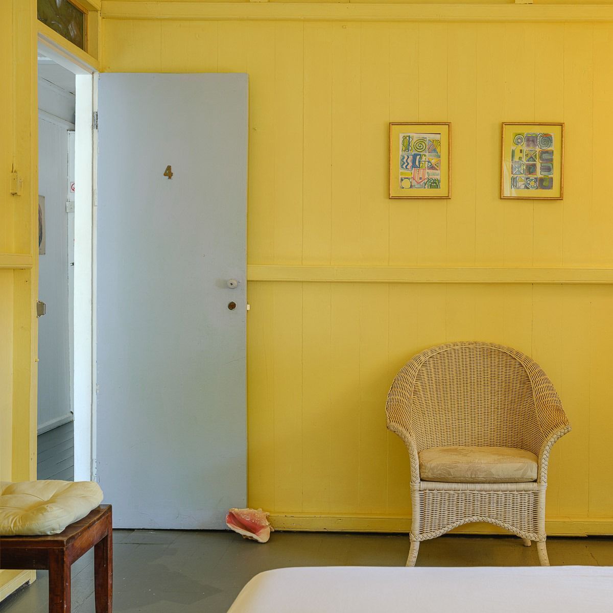 Quarto com parede amarela, porta branca, cadeira de vime e duas obras de arte emolduradas.