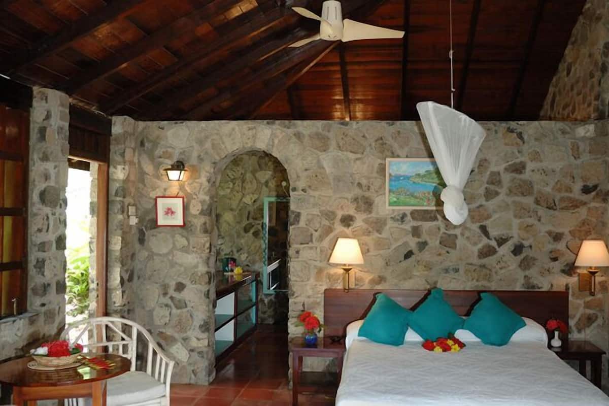 A stone-walled room with a bed, two bedside lamps, a ceiling fan, and a view of the outdoors through an arched doorway.