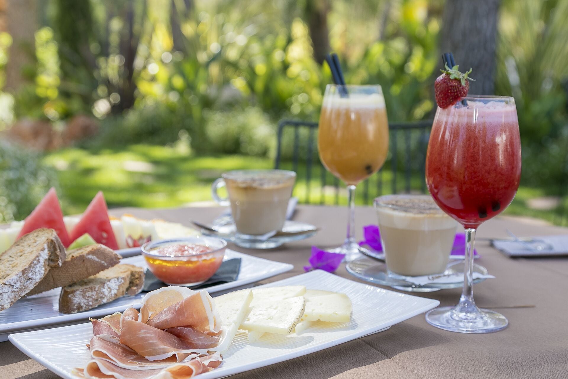 Een buitentafel gedekt met eten en drinken, waaronder parmaham, kaas, brood en twee drankjes in glazen.