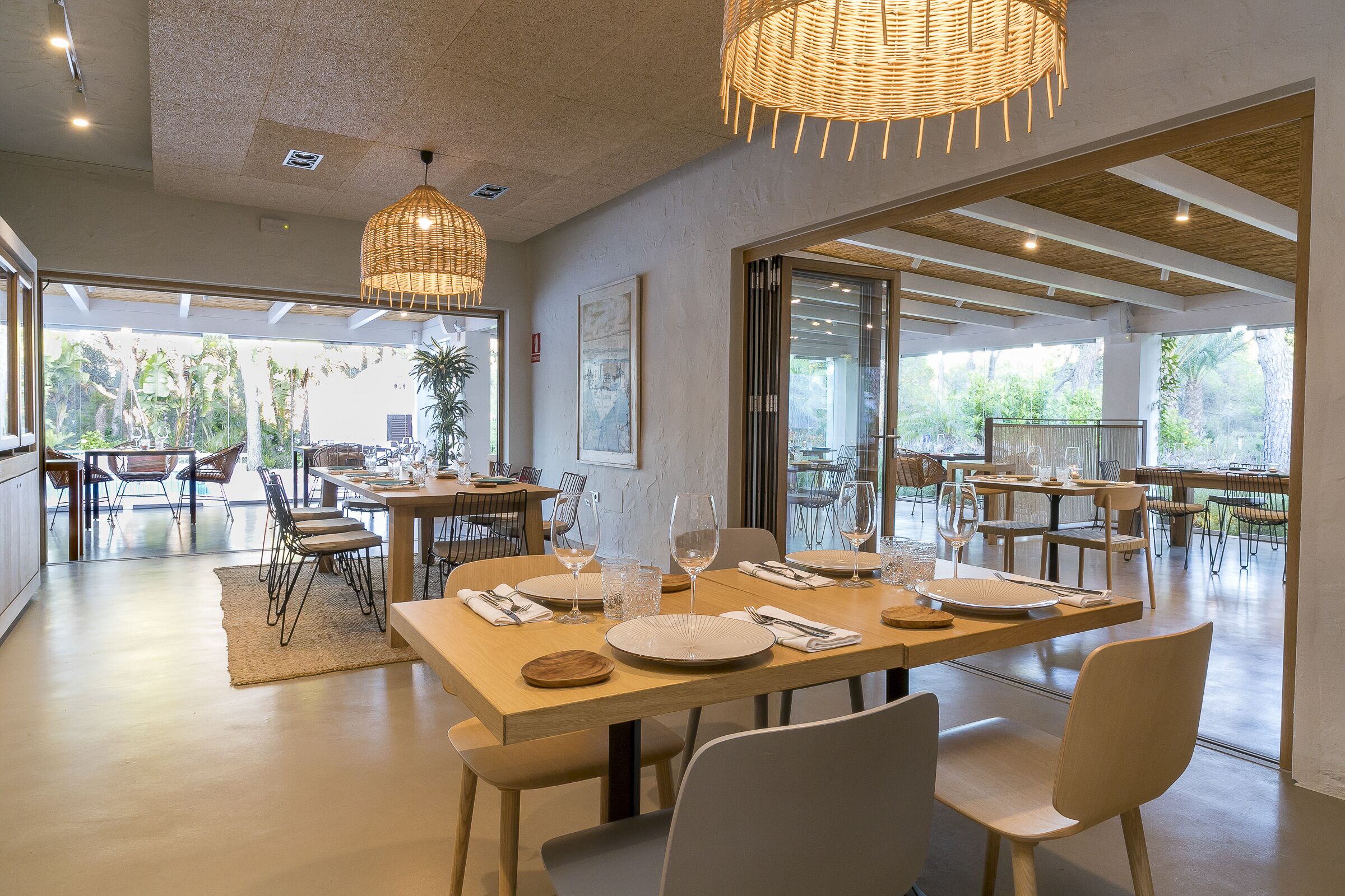 A spacious dining area with wooden tables and chairs, large windows offering a view of the outdoors, and hanging wicker lamps.