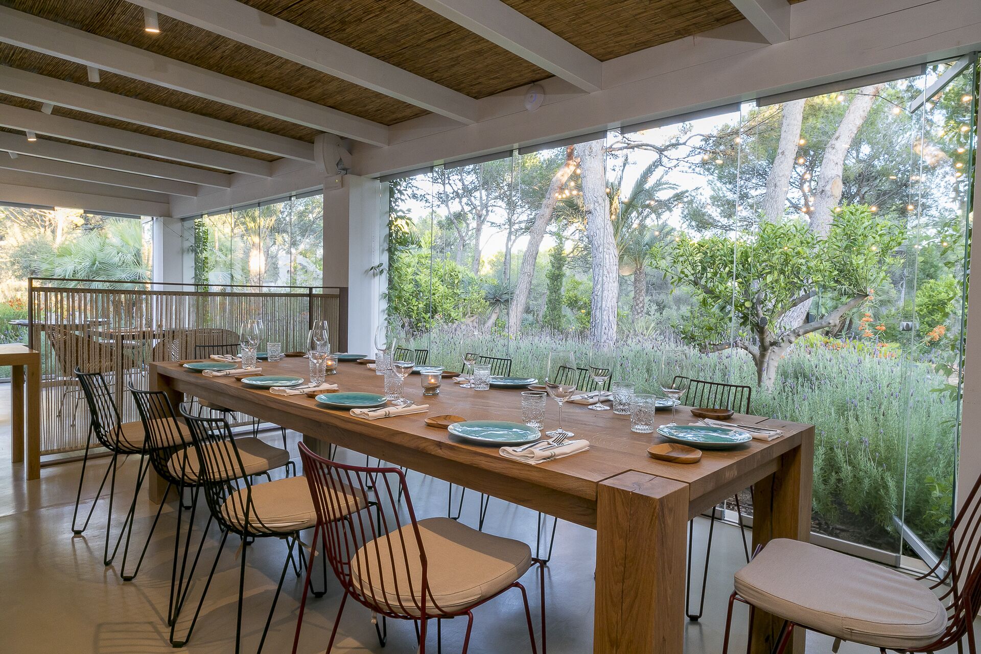 A dining area with a long wooden table set for six, surrounded by chairs, with a view of trees and greenery through large windows.