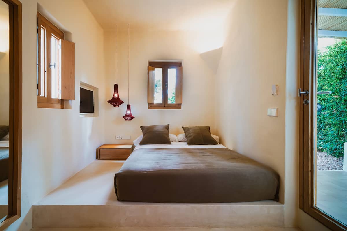 Quarto com uma cama grande, uma janela pequena e vista para o verde lá fora.