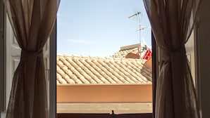 Een slaapkamer met een bed, gordijnen en uitzicht op een pannendak en schoorsteen.