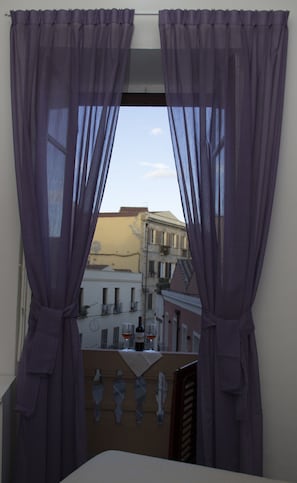 A window with purple curtains, a balcony with a table, and buildings in the background.