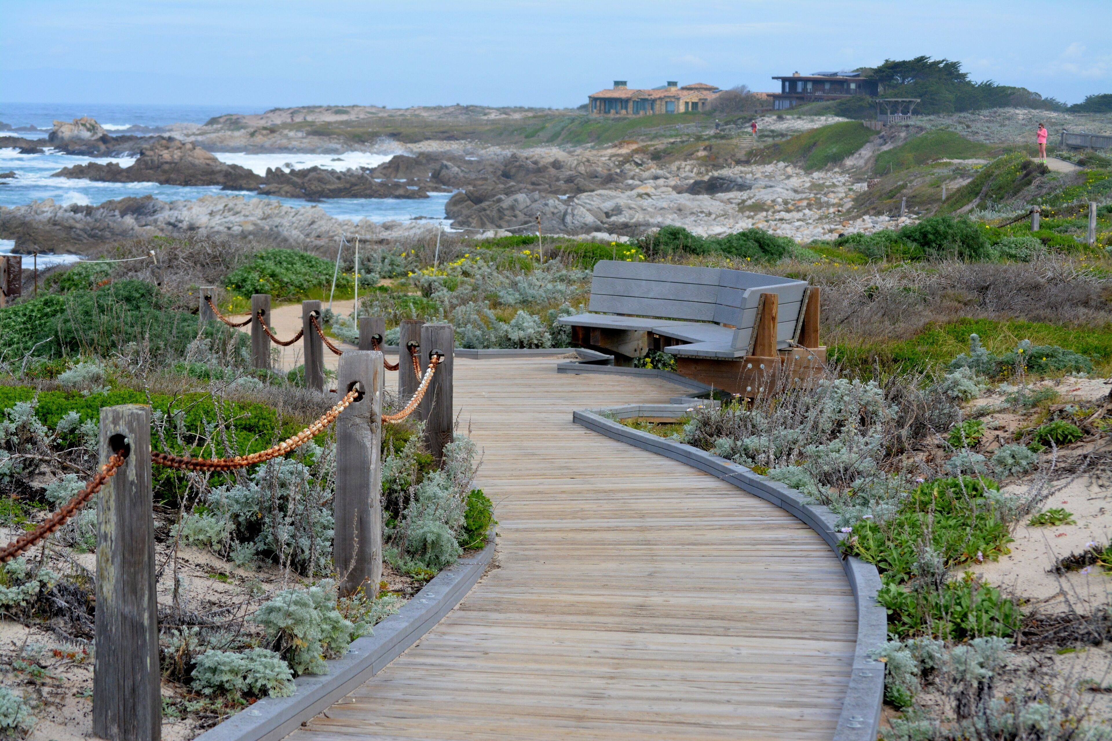 A wooden boardwalk leading to a bench overlooking a coastal landscape with rocky terrain and a building in the distance.