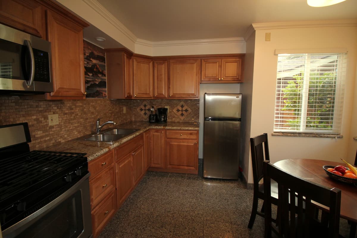 A kitchen with wooden cabinets, a stainless steel refrigerator, a stove, and a dining table with a fruit bowl.