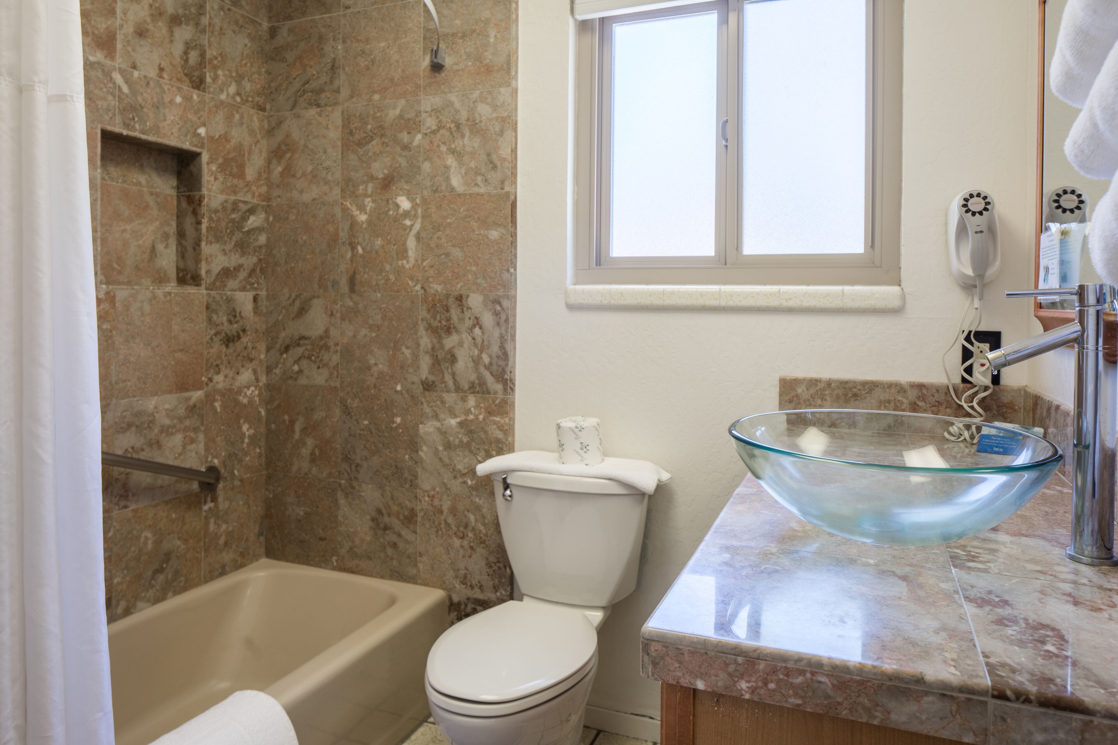 A bathroom with a bathtub, toilet, and a glass bowl sink on a marble countertop.