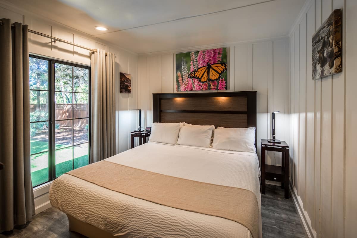 A bedroom with a large bed, a window with curtains, a nightstand with a lamp, and a butterfly painting on the wall.