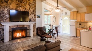 A spacious living room with a stone fireplace, a flat-screen TV, a comfortable brown sofa, and a dining area with wooden furniture.
