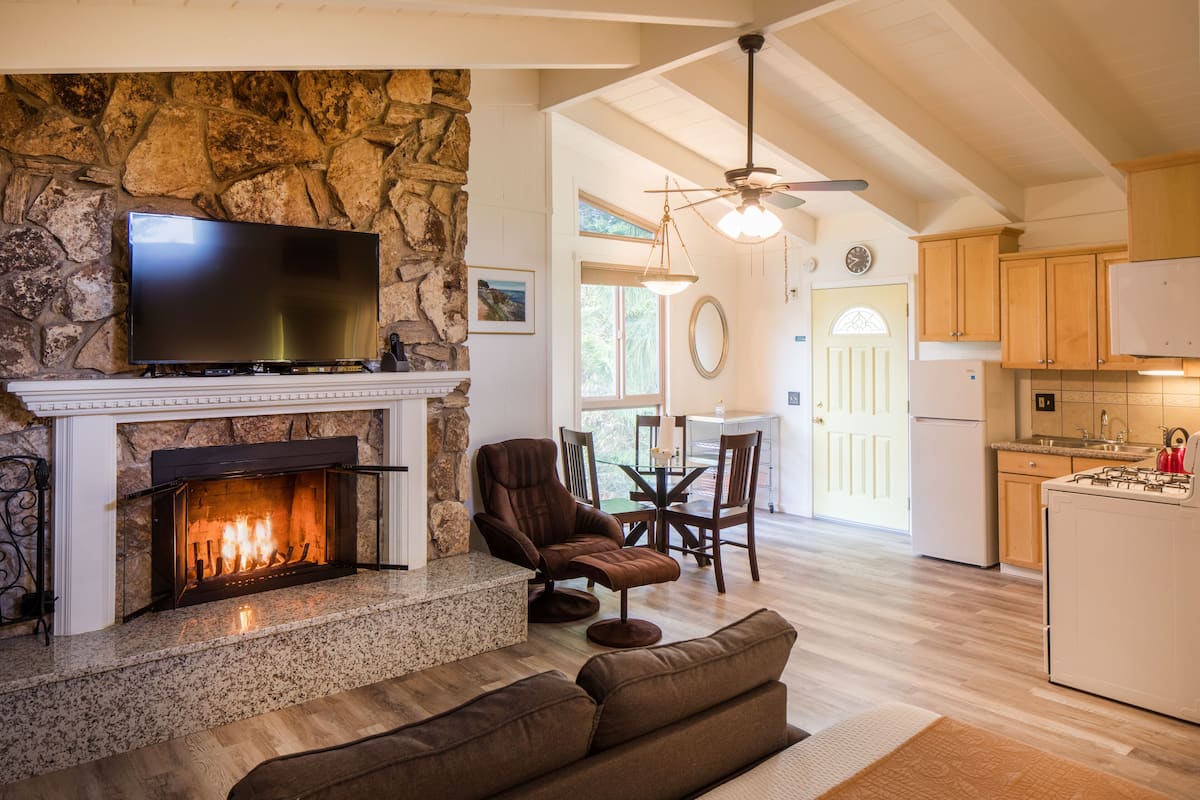 A spacious living room with a stone fireplace, a flat-screen TV, a comfortable brown sofa, and a dining area with wooden furniture.