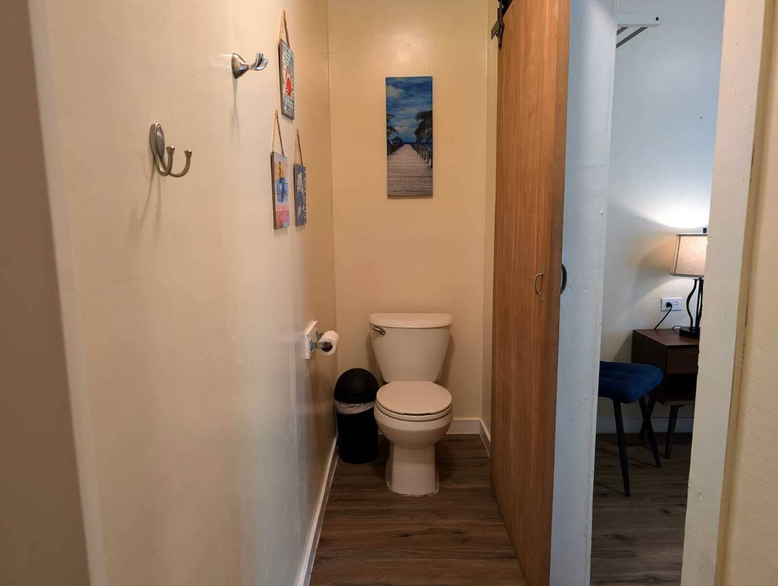 Een smalle gang die leidt naar een badkamer met een toilet, een prullenbak en een klein tafeltje met een lamp.