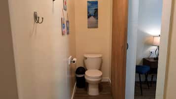Een smalle gang die leidt naar een badkamer met een toilet, een prullenbak en een klein tafeltje met een lamp.