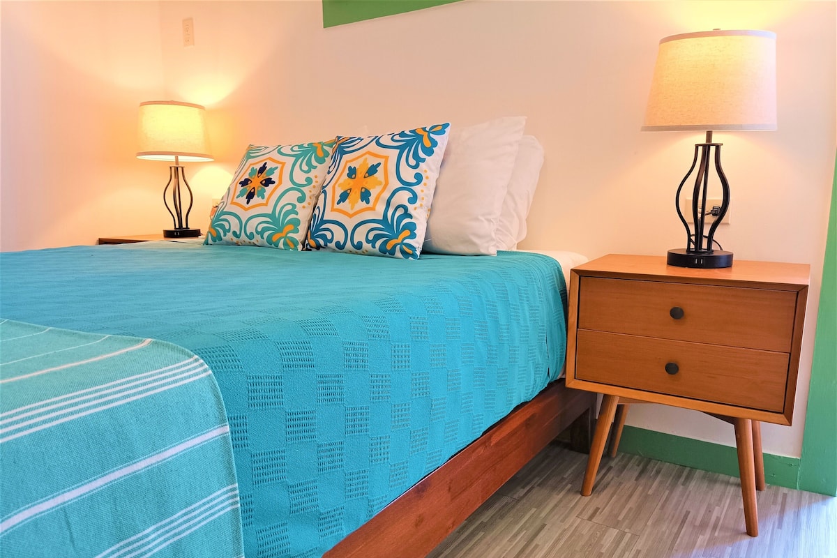 A neatly made bed with a blue blanket and patterned pillows, a wooden nightstand with a drawer, and two wall-mounted lamps.