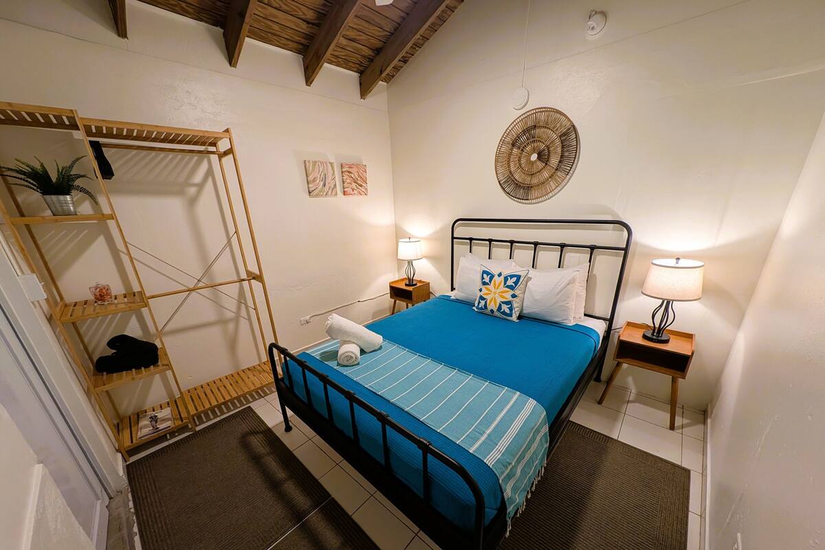 A bedroom with a bed, two bedside tables, a wall-mounted fan, a shelf with a plant, and a door with a towel on it.