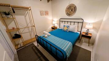 A bedroom with a bed, two bedside tables, a wall-mounted fan, a shelf with a plant, and a door with a towel on it.