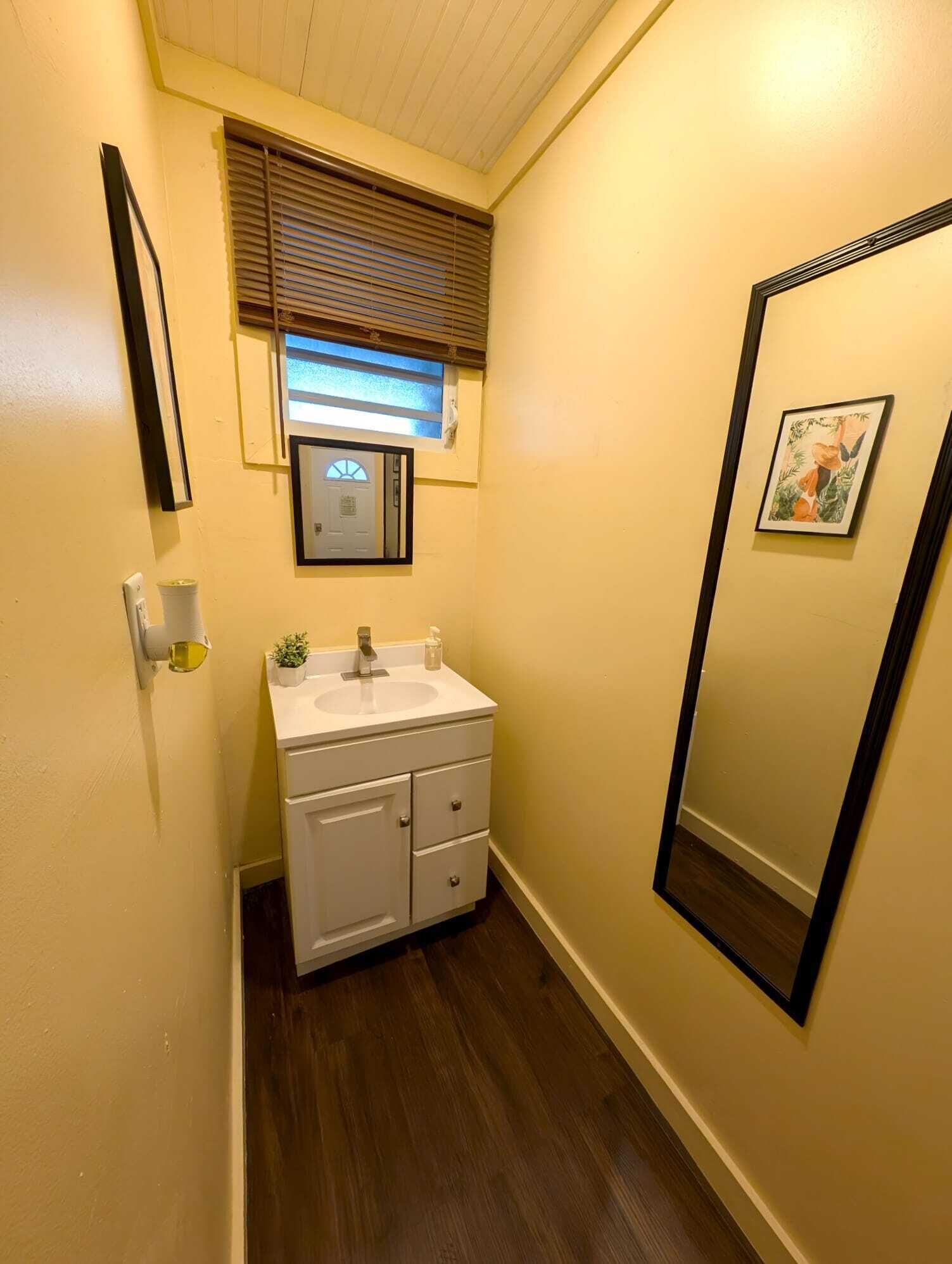 A narrow bathroom with a white sink, a mirror, and a window with blinds.
