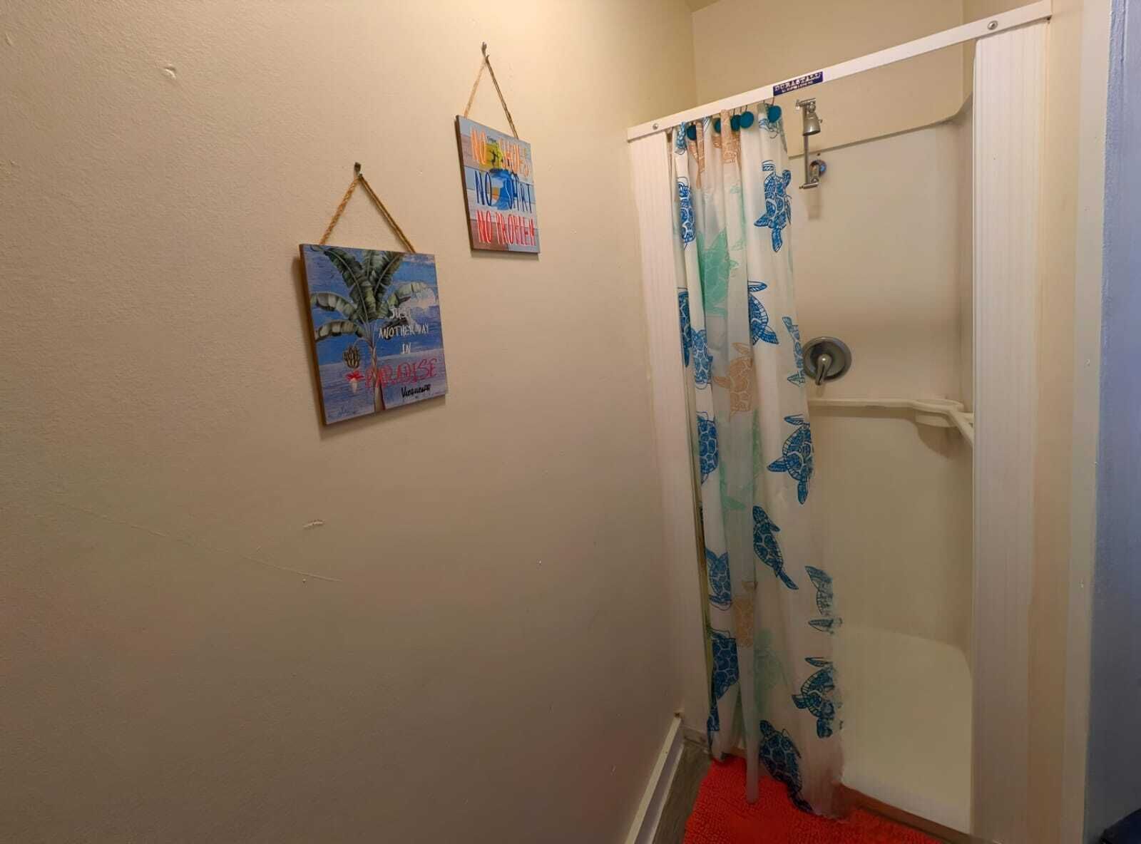 Une salle de bain avec un rideau de douche orné d’un motif de tortue de mer, un porte-serviettes avec deux serviettes et deux tableaux accrochés au mur.
