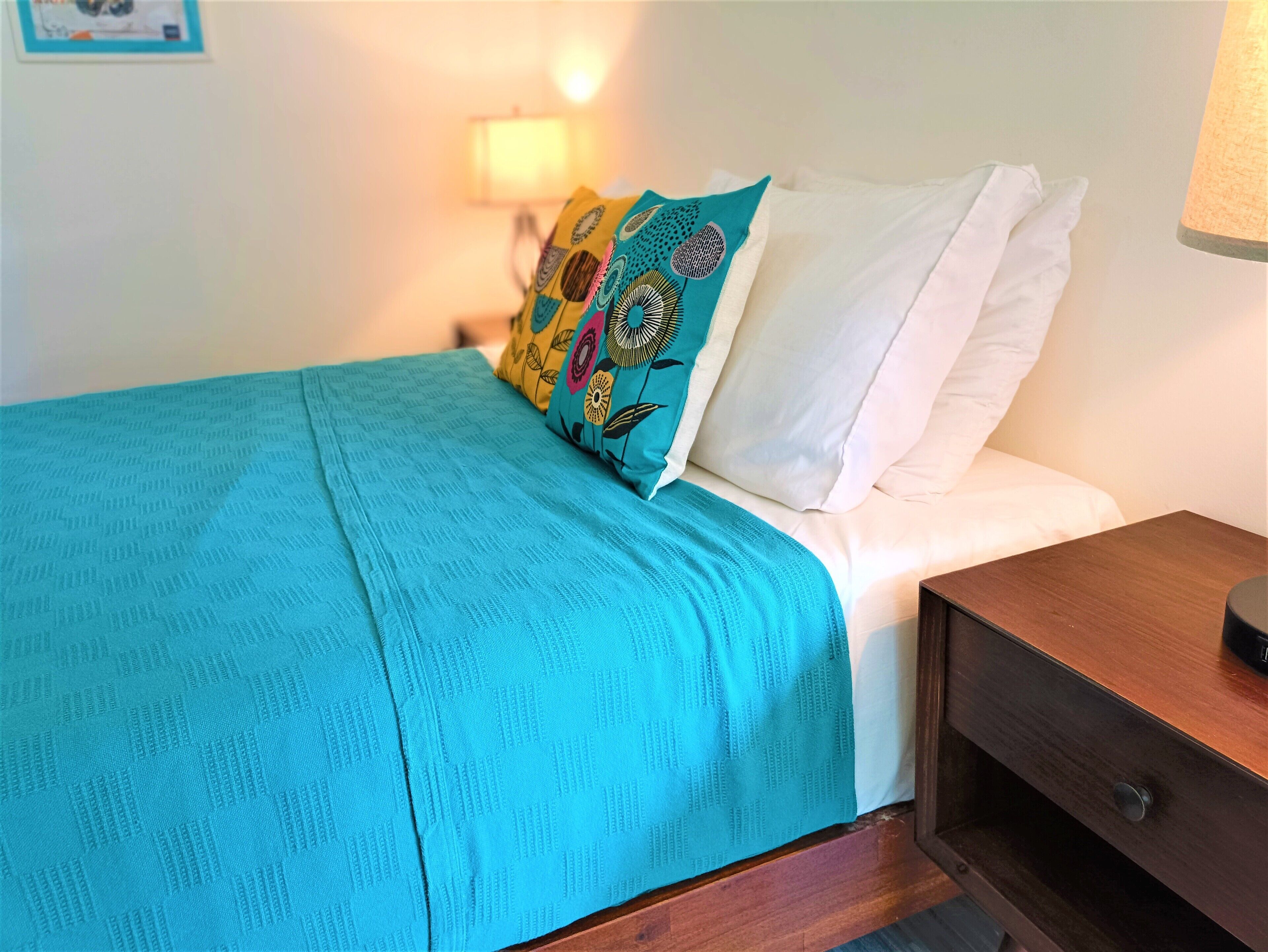 Un lit avec une couverture bleue et un oreiller coloré, un autre oreiller blanc, et une table de chevet en bois avec une lampe.