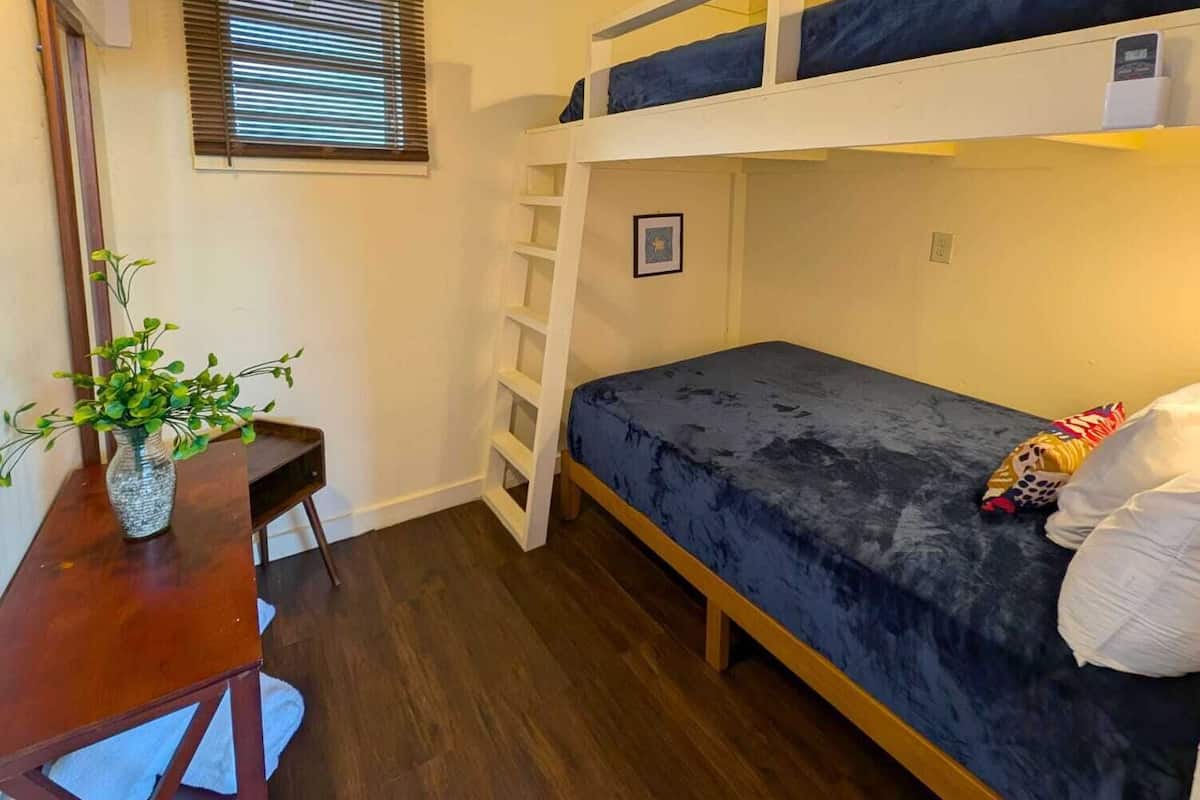 A bunk bed room with a desk and chair, a small table with a vase, and a window with blinds.