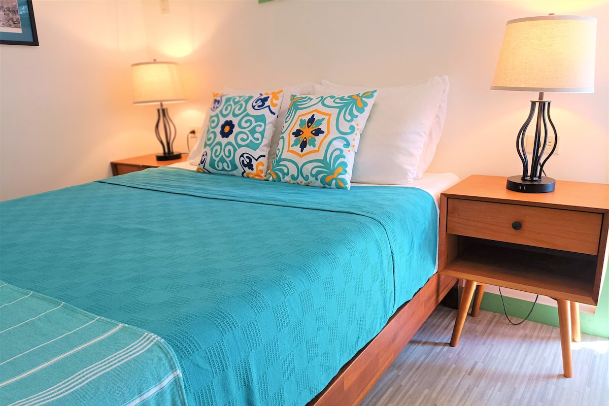 A neatly made bed with a teal bedspread and patterned pillows, flanked by two bedside tables with lamps, one on each side.