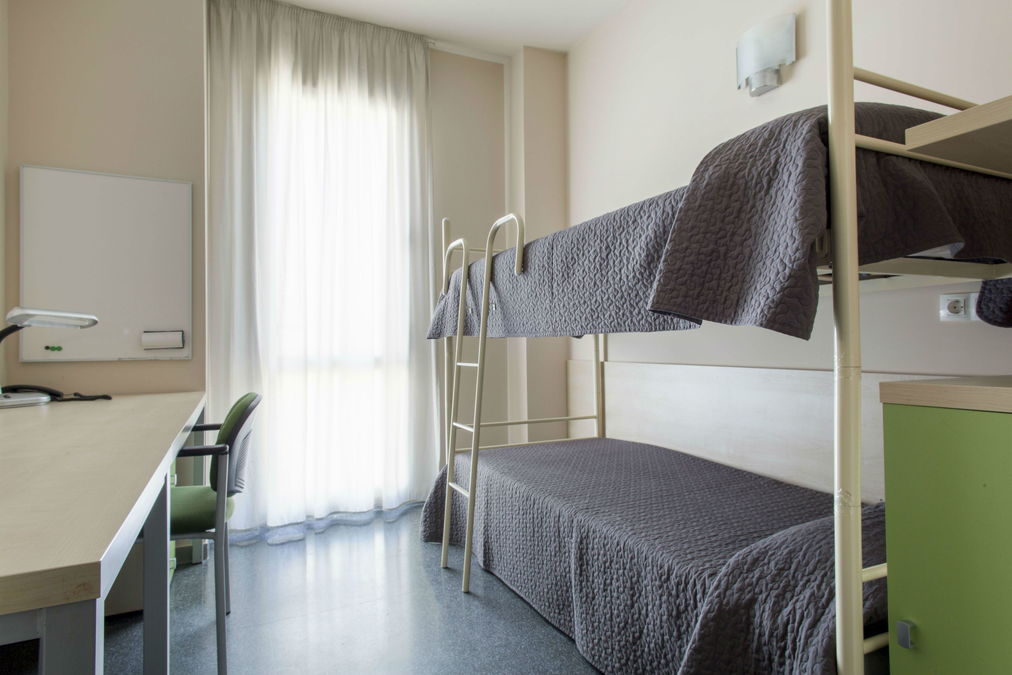 Een stapelbed met een bureau en stoel, een raam met gordijnen, en een groene kast.