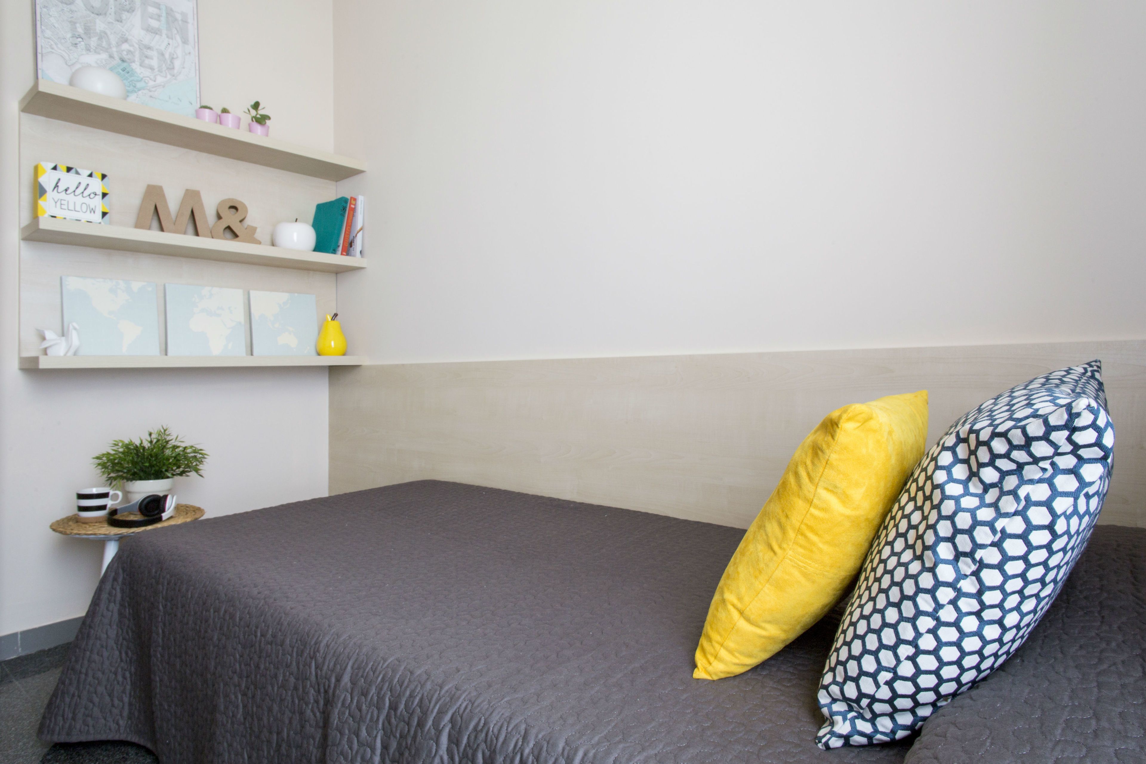 Un dormitorio ordenado con una cama gris, dos almohadas, una mesita de noche con una planta y una estantería con objetos decorativos.