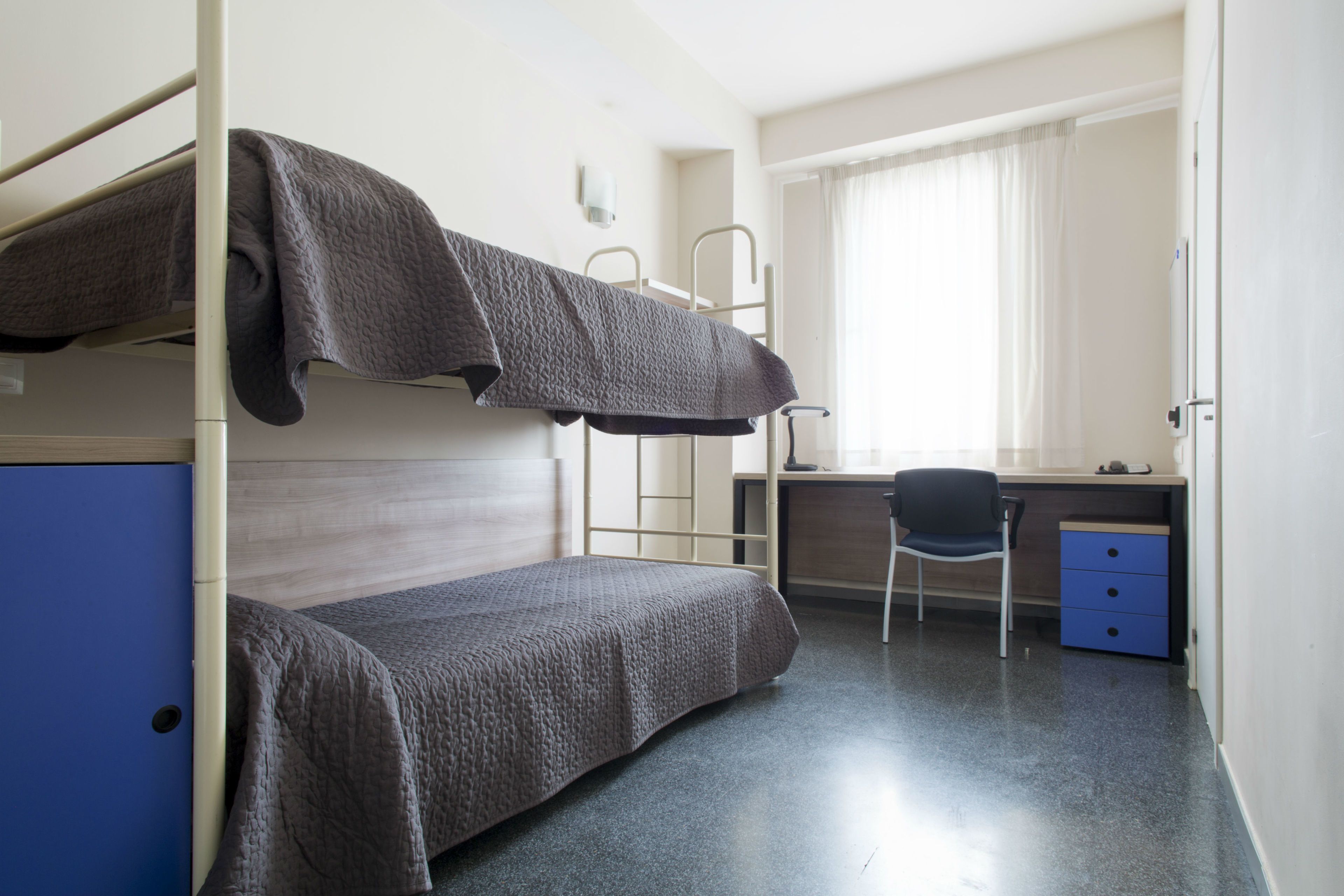 Una cama litera con escritorio y silla, un armario pequeño y una ventana con cortinas.