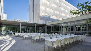Un espacio de comedor al aire libre con mesas y sillas, un corredor cubierto y un edificio moderno al fondo.