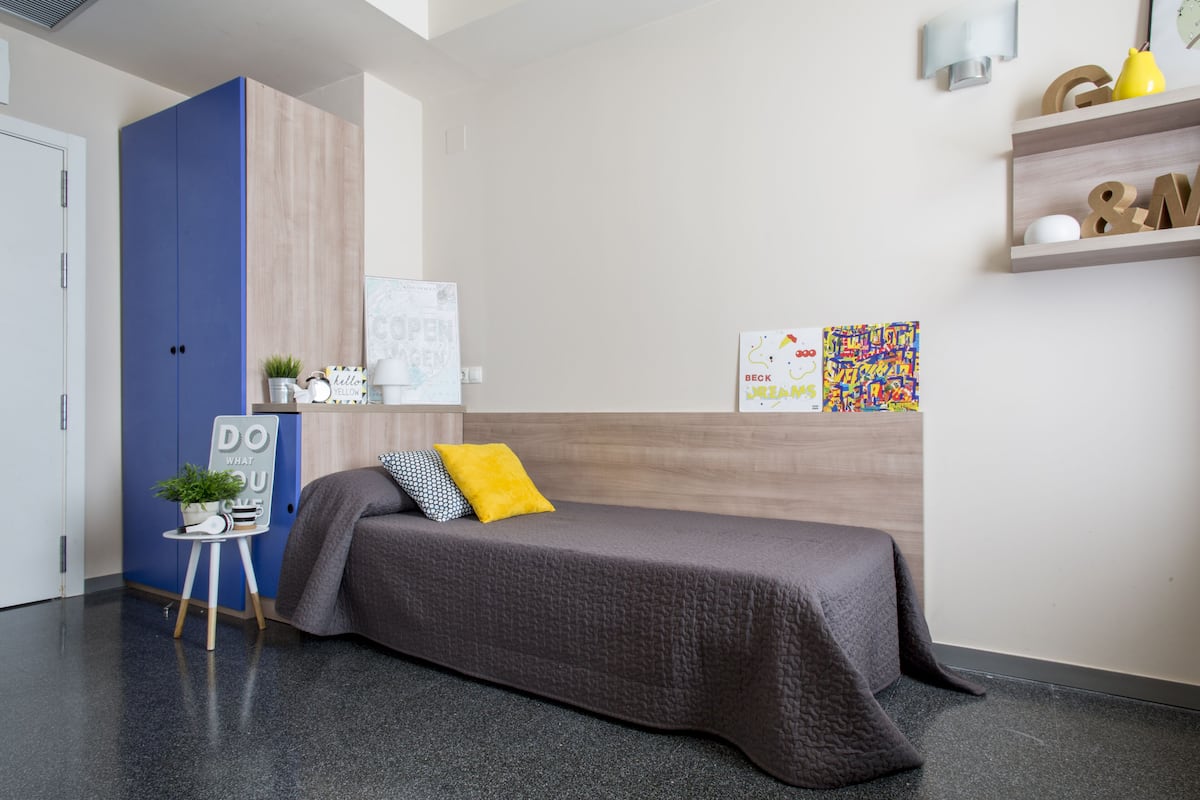 Et moderne soveværelse med en seng, en blå garderobe, et lille skrivebord med en plante og en hylde med dekorative genstande.
