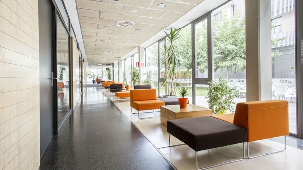Un espacio interior moderno con un largo pasillo bordeado de asientos naranja y negro, ventanas amplias y plantas en macetas.