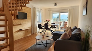 Een moderne woonkamer met een houten trap, een flatscreen televisie bevestigd aan een houten wand, een eethoek met stoelen en een tafel, en een bank met kussens.