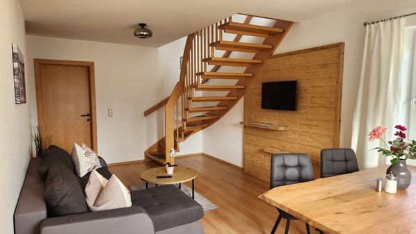 A modern living room with a wooden staircase, a flat-screen TV mounted on a wooden wall, a dining table with chairs, and a sofa with cushions. - Pension Ballwein (Salzburg)