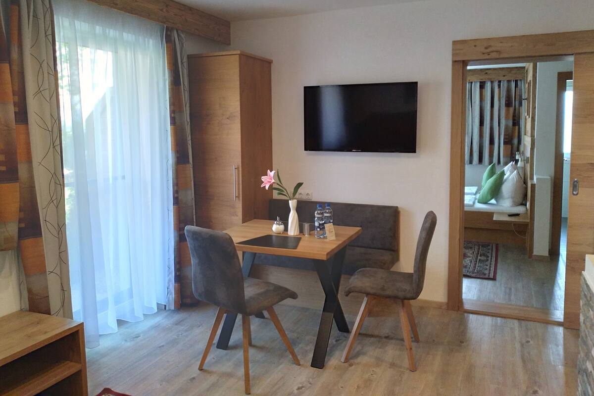 Un salon moderne avec une table à manger en bois, deux fauteuils gris à revêtement, un téléviseur à écran plat fixé au mur et une armoire en bois.