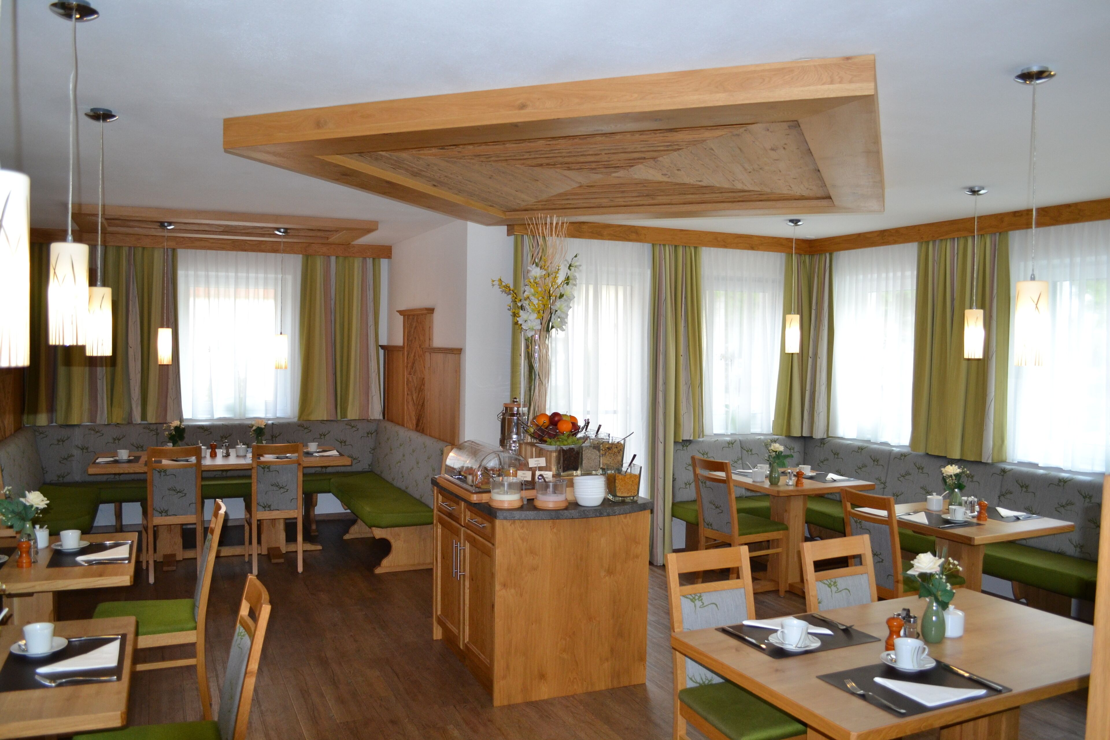 Ein Restaurant mit Holztischen und grün gepolsterten Sitzplätzen, das eine zentrale Insellounge mit einer Blumenvase zeigt.