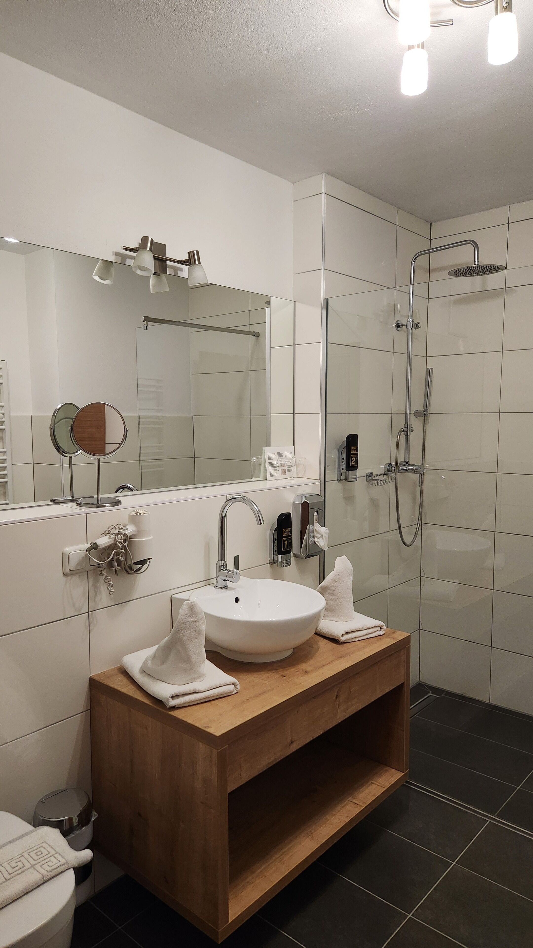 Un baño moderno con tocador de madera, un espejo grande, un lavabo blanco y una ducha con cerramiento de vidrio.