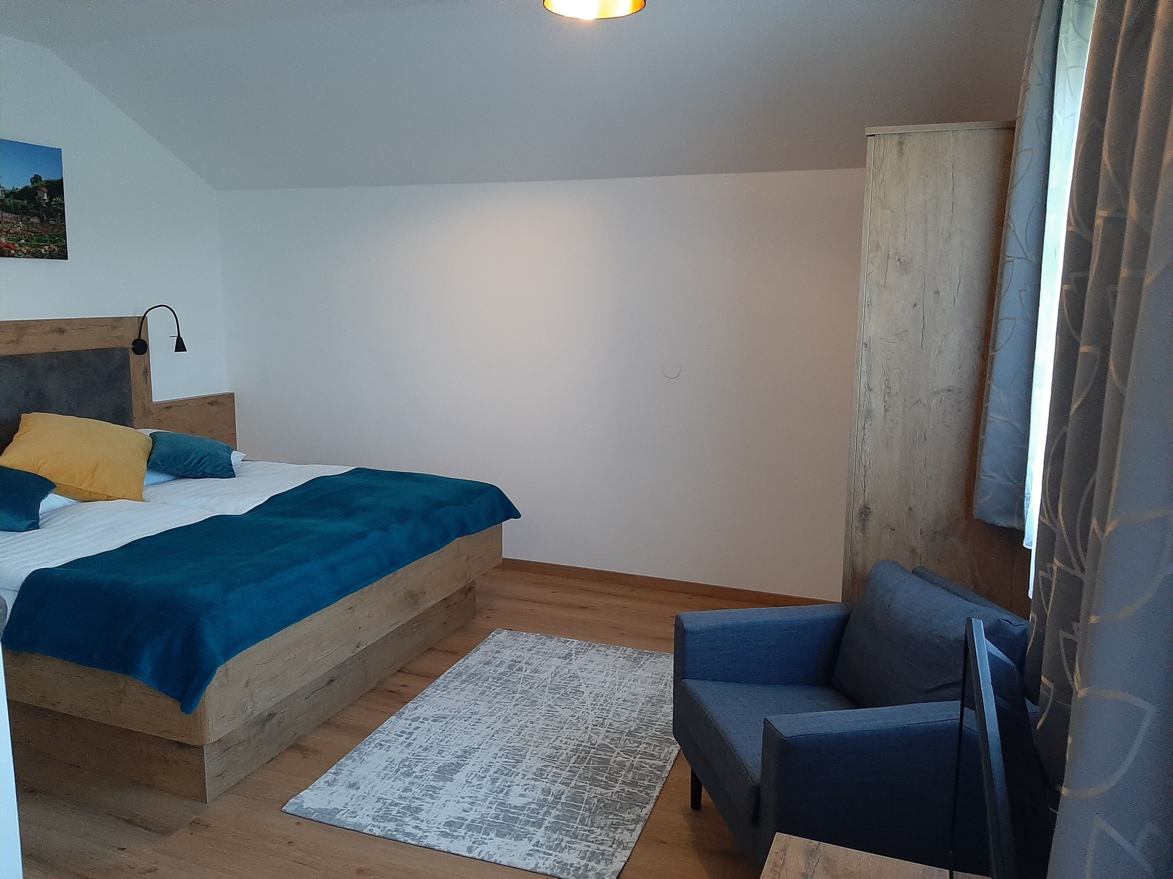 A bedroom with a wooden bed, a blue chair, a small rug, and a wall-mounted lamp.