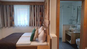 Ein Schlafzimmer mit einem Holz-Kopfteil, einem Bett mit weißen Leinen und Blick auf ein Badezimmer mit Dusche und Waschbecken.
